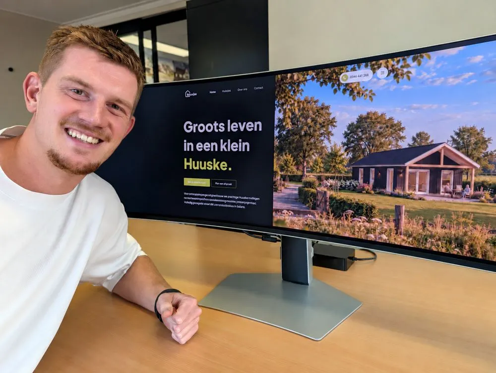 Een lachende man bij een brede monitor met de website van Huuske